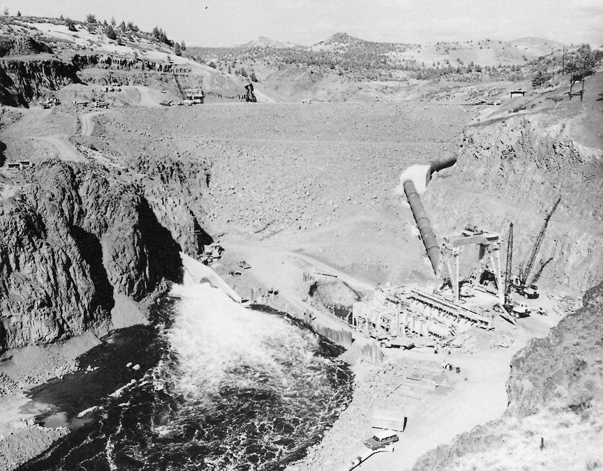 Old black and white photograph of Iron Gate dam construction site on the Klamath River. Surrounding the river is a dug-out rocky basin and large construction machinery. 