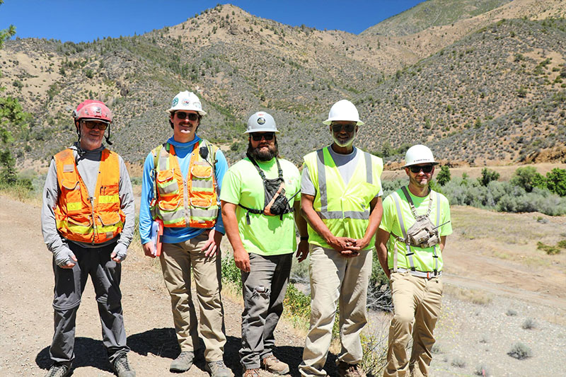 5 men outdoors in safety gear posing for a photo with mountains in the background